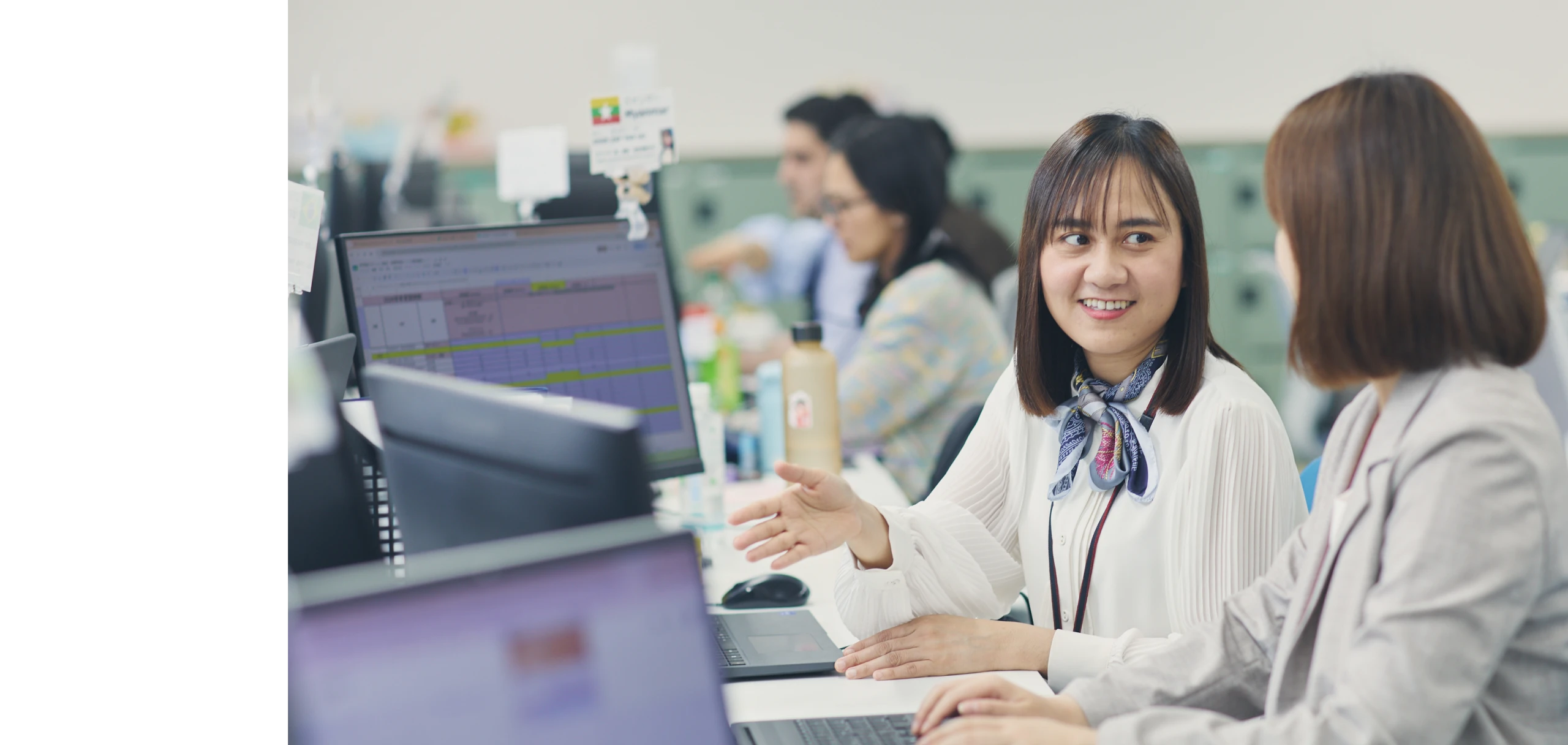 オフィスで同僚と笑顔で話しながらパソコンの画面を指し示す女性社員