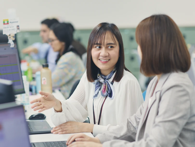 オフィスで同僚と笑顔で話しながらパソコンの画面を指し示す女性社員