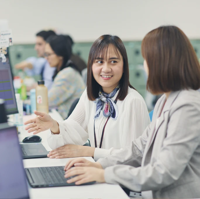 オフィスで同僚と笑顔で話しながらパソコンの画面を指し示す女性社員